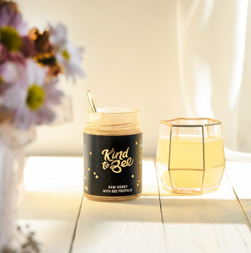 A see through glass of water mixed with honey with propolis standing next to the jar of honey and a bouquet of flowers